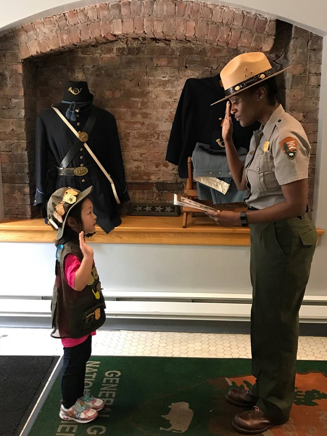 Junior ranger on the left wearing a vest and hat, holding right hand up while on the right, a Park Ranger in uniform holds their right hand up while reading from junior ranger booklet in left hand.