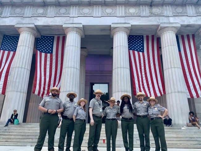 8 Park Rangers standing side by side wearing their green and grey uniforms, while the mausoleum is in the background with American flags hanging down