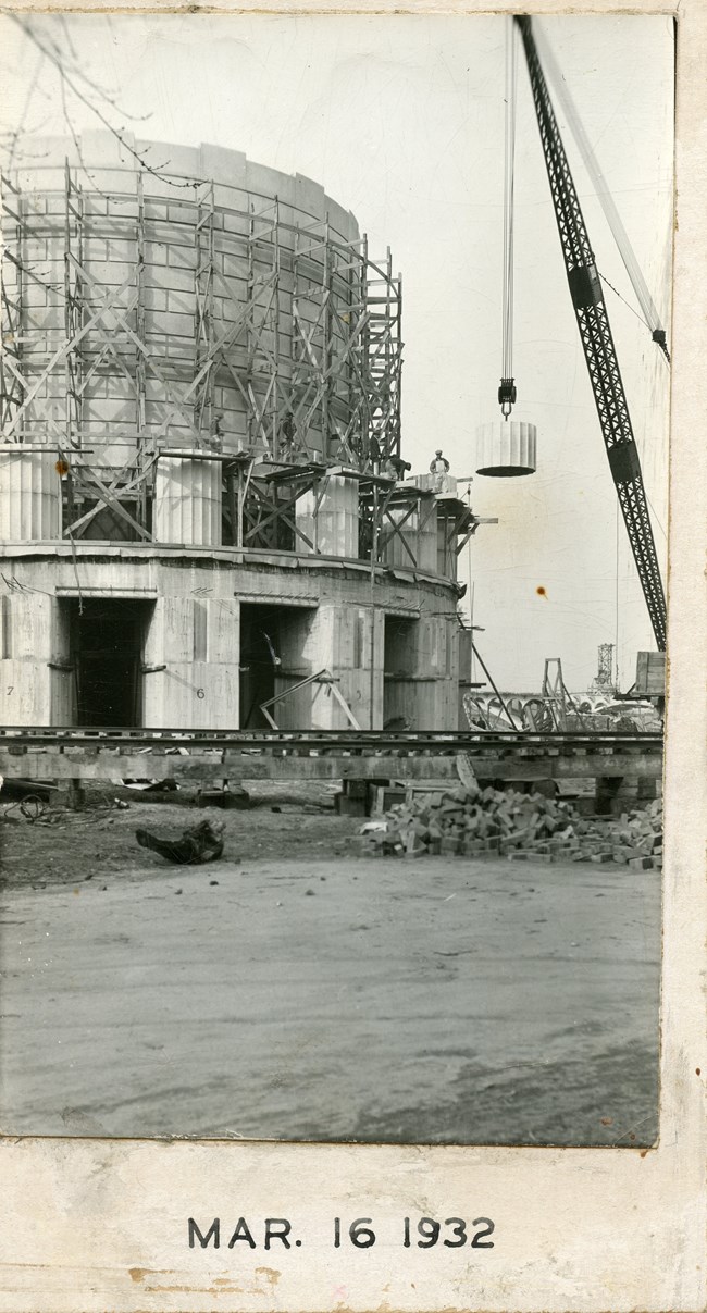 Part of the memorial can be seen, the first section of the doric columns have been placed a crane lifts another drum of a column into place. Construction materials and a railroad track are on the ground in front of the memorial.