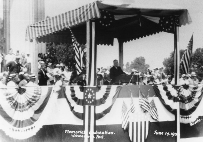 A black and white photo of President Franklin Delano Roosevelt stands behind a podium on a stage built on the memorial steps decorated with American flags and draped with stars and stripes cloth. A striped awning covers the podium area.