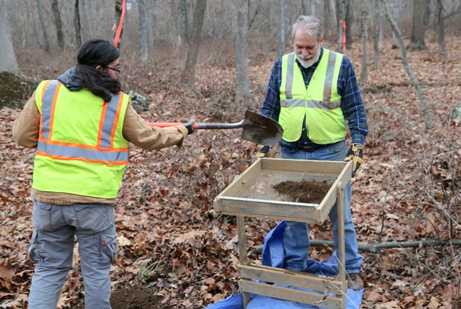 On the left, a woman uses a shovel to throw dirt into wooden-framed screen. On the right, a man shakes the dirt through the screen to look for artifacts. They are in the woods and both are wearing bright green safety vests.