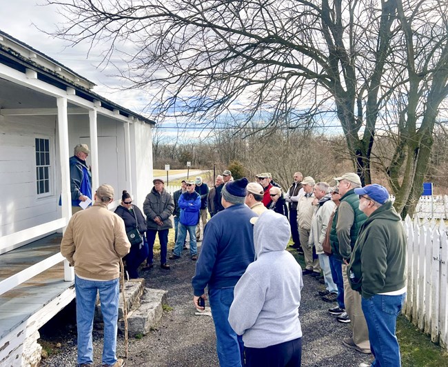 A large crowd gathers outside a small white historic house. On the porch stands a man that the crowd of people is listening to. To the right of the crowd is a white picket fence and to the right is a tall bare tree.
