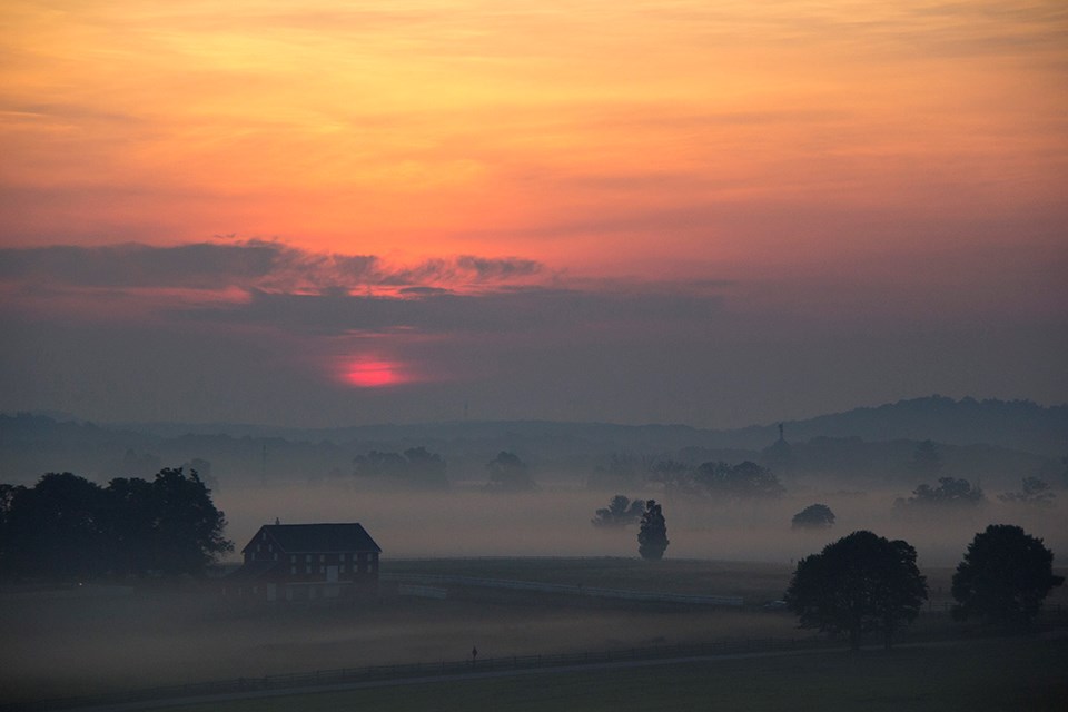 A sunrise on a foggy morning. There is fog near the ground, a barn in the lower left corner, trees dot the landscape, and the sun is barely peaking through the clouds with a soft yellow and orange sky.
