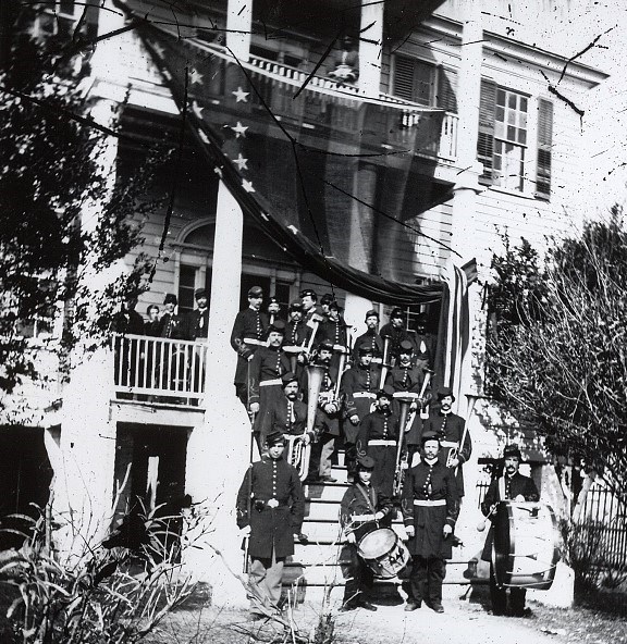 Musicians of a Civil War band stand on the stairs of a large house