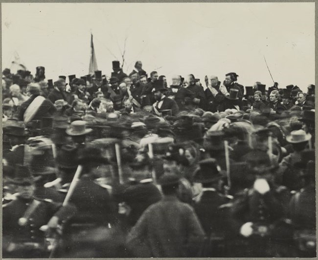 A large crowd gathers around a platform upon which is Abraham Lincoln and a host of other dignitaries in attendance for the dedication of the Gettysburg Soldiers' National Cemetery