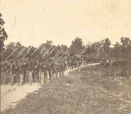 Photograph showing Civil War Soldiers of the 134th Illinois Infantry On The March; the soldiers are carrying rifles with bayonets attached.