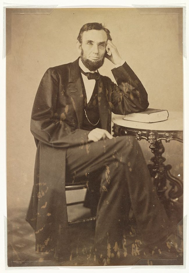 A photograph of a seated Abraham Lincoln at table with left hand held against his head, looking into camera.