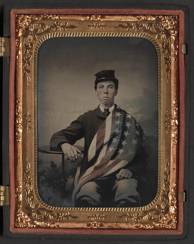 Photograph showing an unidentified U.S. Army Civil War soldier with an American flag draped over his right shoulder.