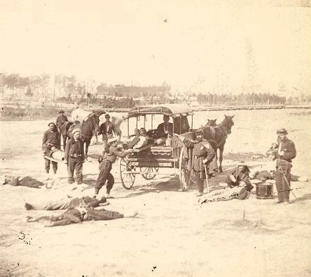 Stereograph showing members of the Ambulance Corps carrying wounded soldiers on stretchers to the horse drawn ambulance in the field.