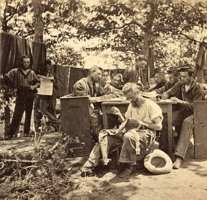 A group of Civil War soldiers sit around a table in camp writing letters while another soldier sews his uniform