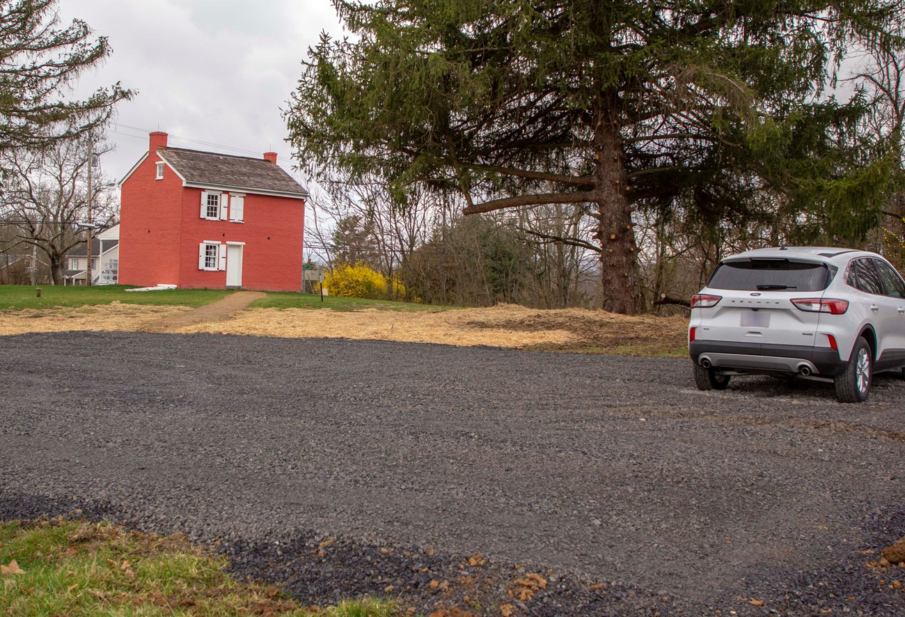 In the foreground is a dark gravel parking lot with a small white SUV on the right. In the distance in the center is a large pine tree and on the left is a small, two story, red brick house with white shutters and door.