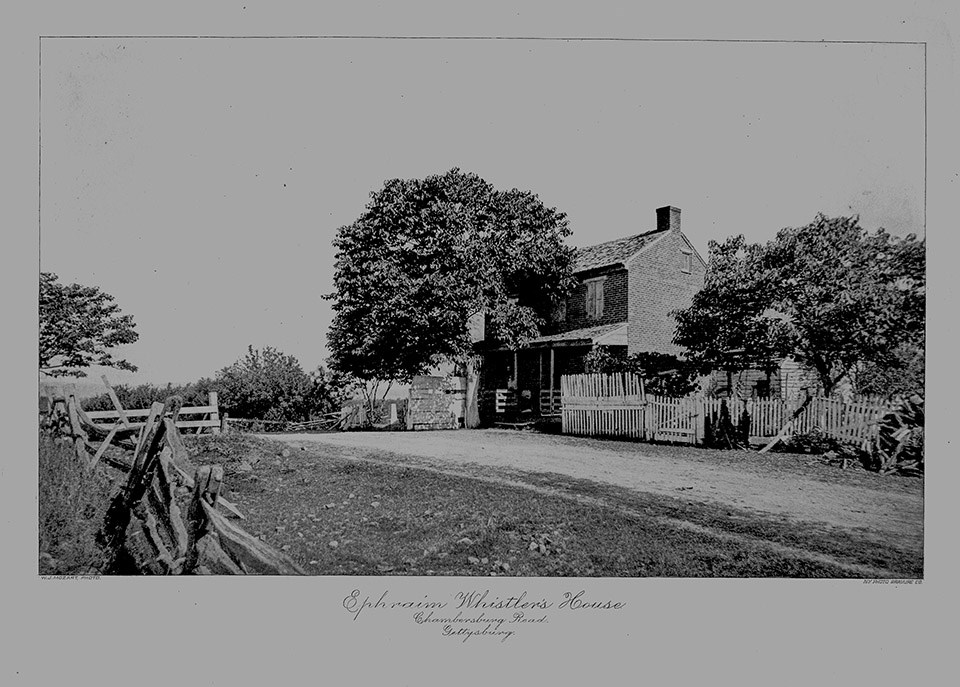 A black and white photograph of a brick house sitting atop a rise of ground surrounded by trees. A white picket fence is in front of the house and is next to a dirt road. Wooden fences line the near side of the road.