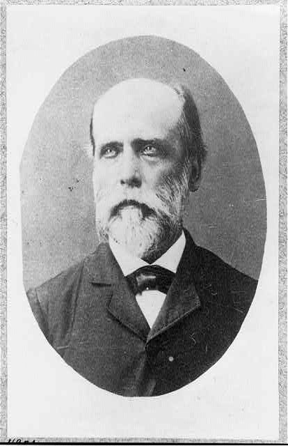 Black and white image of a balding man with a gray beard wearing a black coat