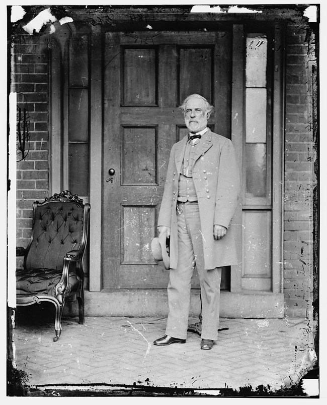 A man in a gray military uniform stands on the porch of a house in front of a door