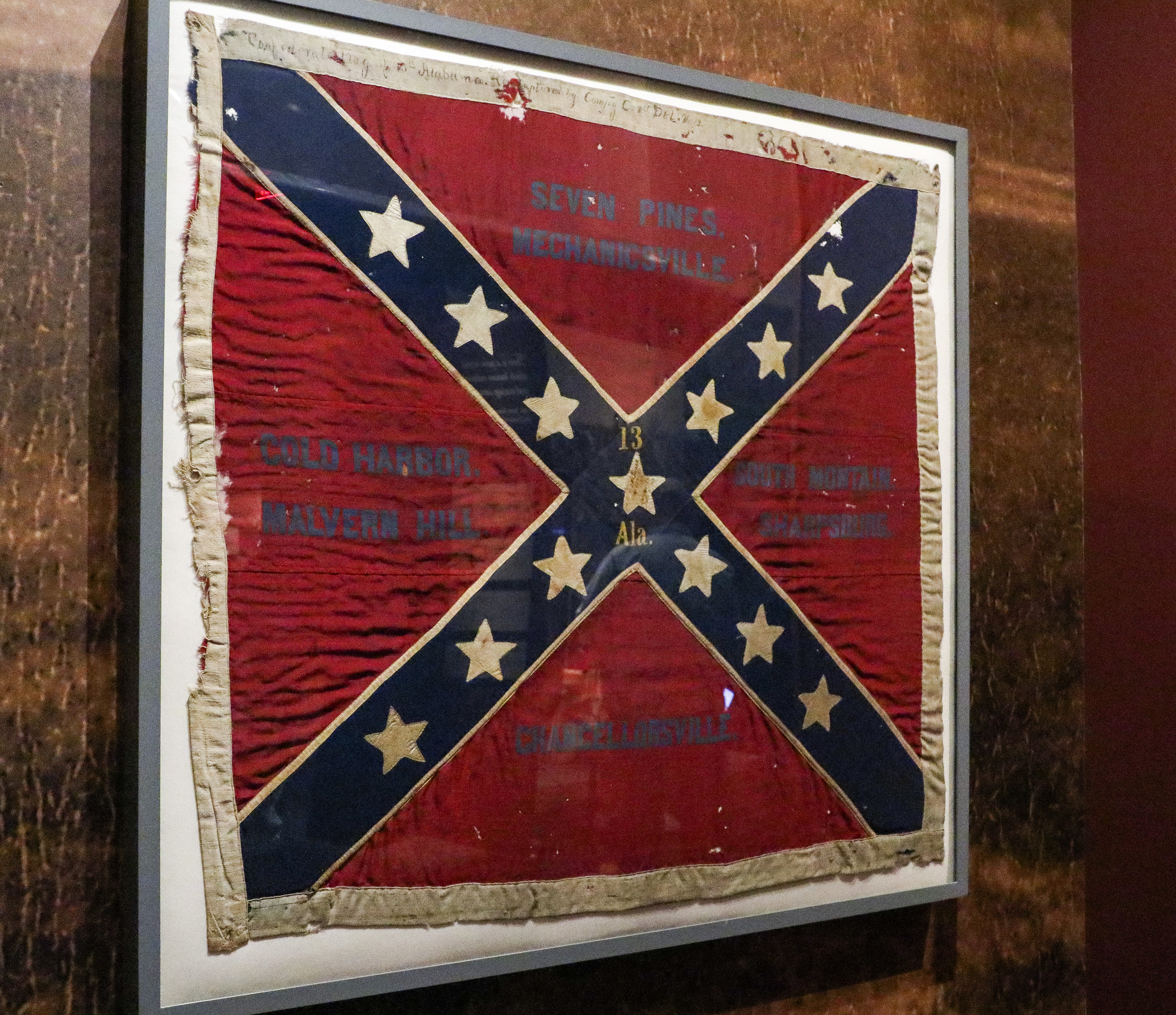 A Confederate battle flag hangs on a wall. The flag has a red background with diagonal blue lines and thirteen white stars on the blue lines.