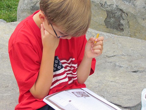 A young boy, dressed in a red t-shirt with an American flag on it, reads his Junior Ranger book and answers questions.