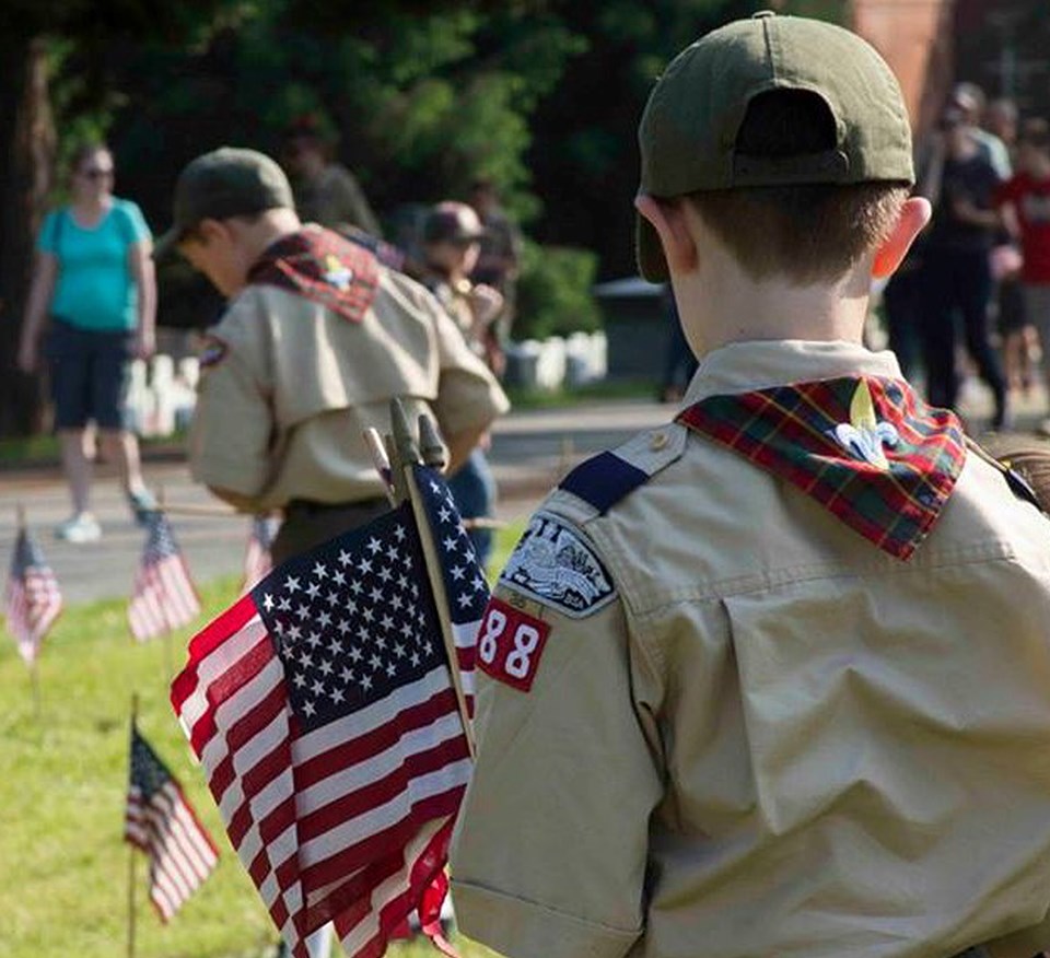A boy scout is seen from behind as he holds a bunch of small American flags.