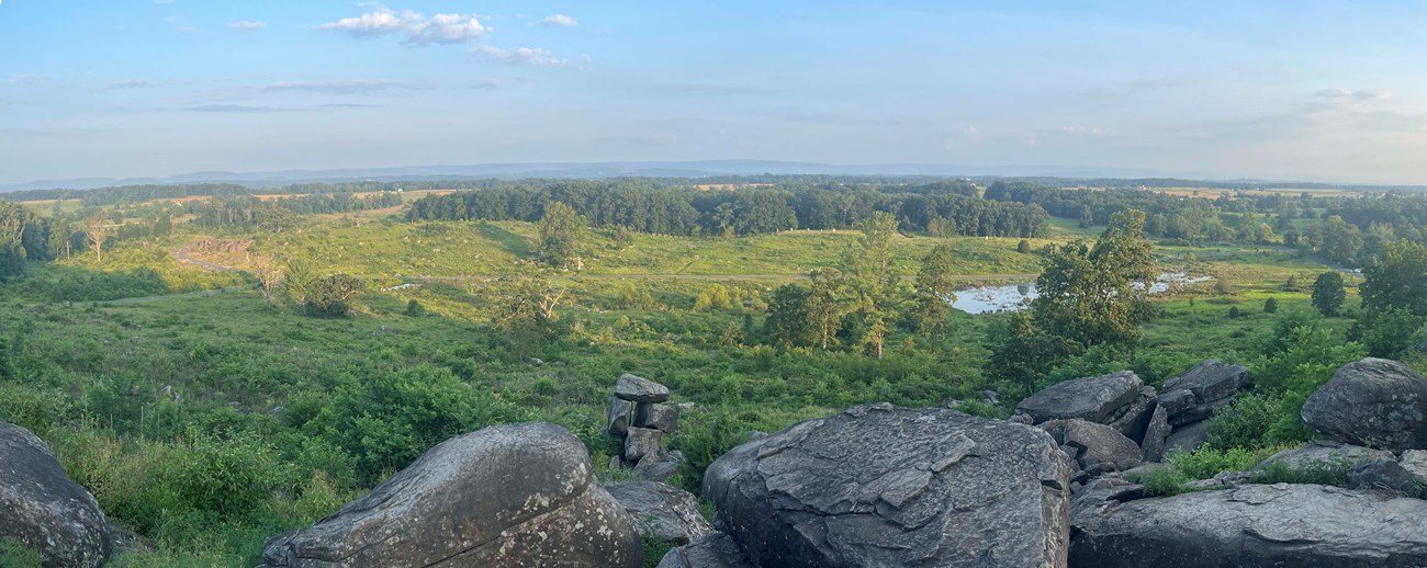 A wide angle view from the summit of a rocky hill looking down into a lush green valley. A pond is on the right and thick trees are nearby. A blue sky with a few white clouds are above.
