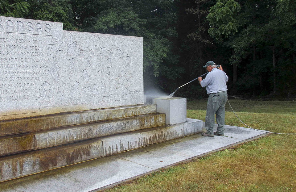 A park employee uses a pressure washer to clean the Arkansas Memorial. There is an etched wall of soldiers and three steps of light colored granite.