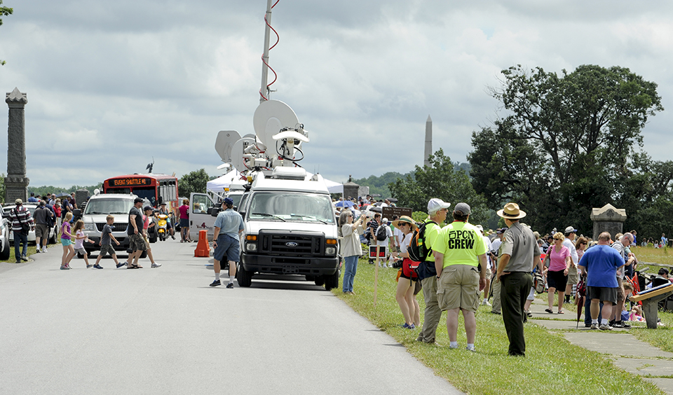 Press trucks are parked along the side of the road. A park ranger talks to visitors in the foreground and lots of visitors walk around the battlefield.