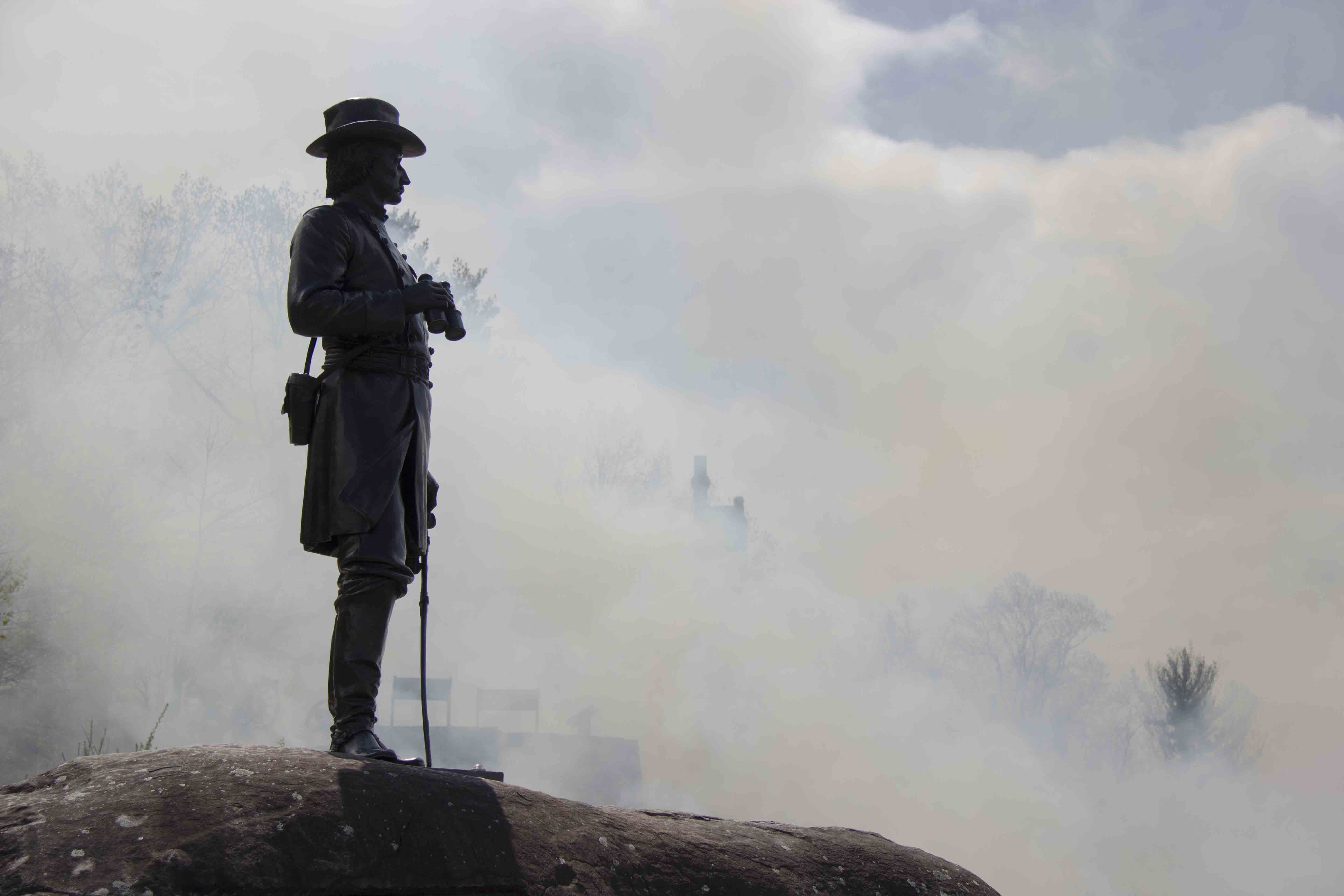 A statue of a soldier stands on top of a boulder as smoke billows up behind it.