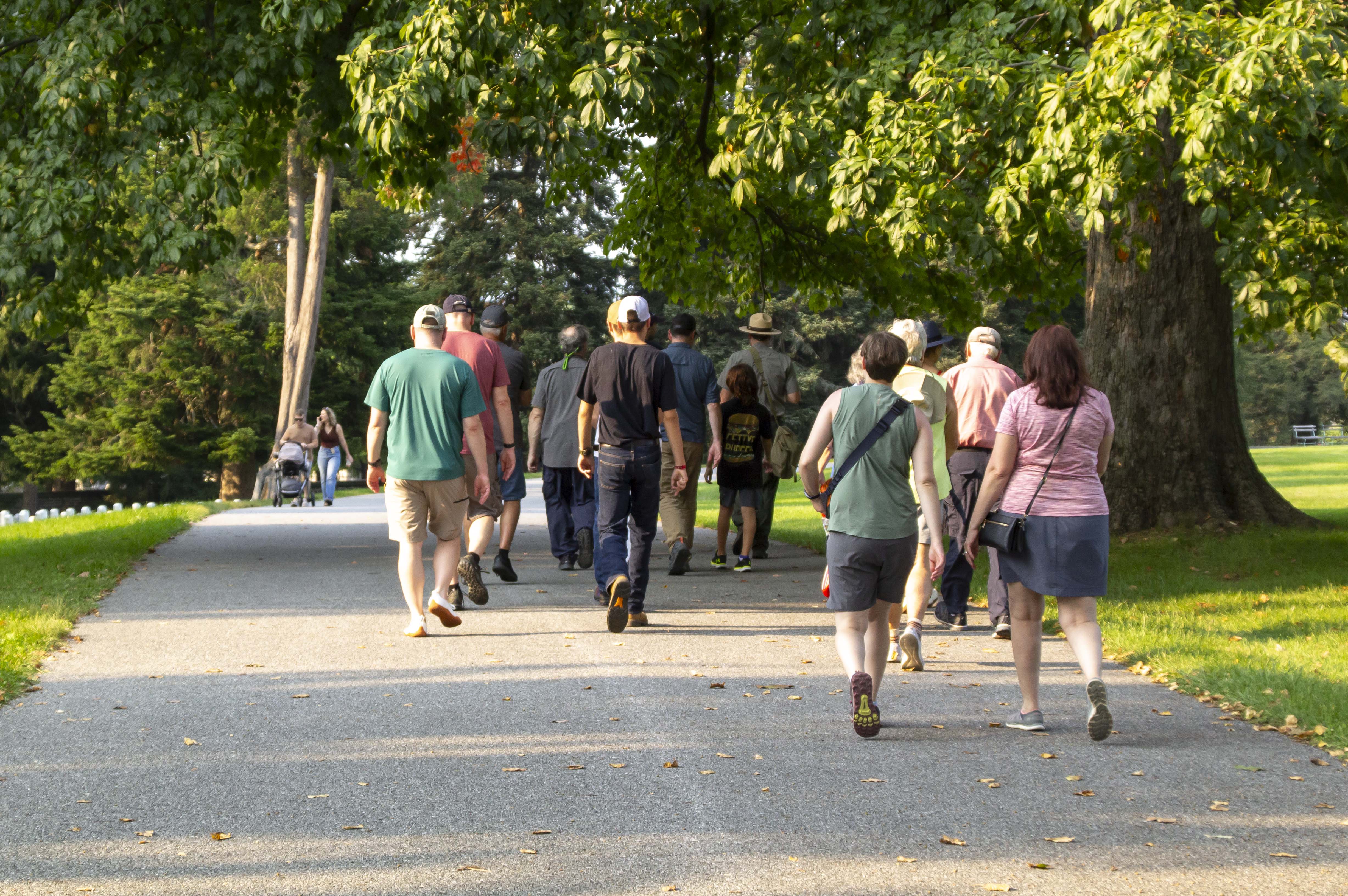 A group of visitors walk away from the camera following a park ranger along a wide paved path into a cemetery.