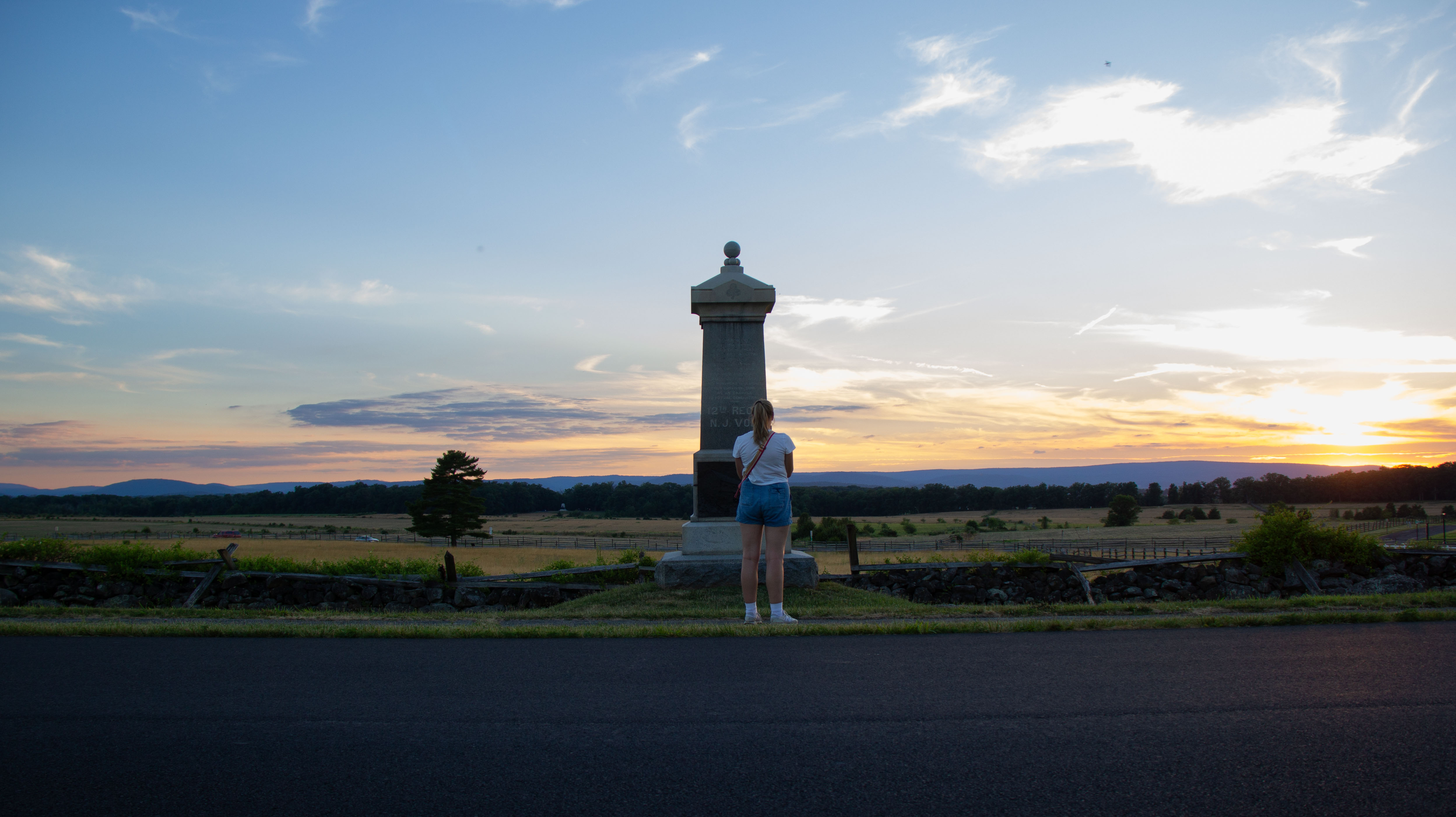 A person stands on a road in front of a stone monument at sunset