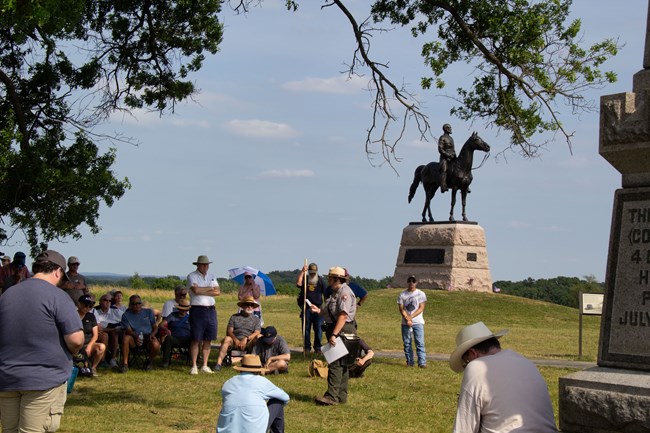 A stone and bronze monument of soldier on a horse stands in a field. A park ranger gives a talk to people.