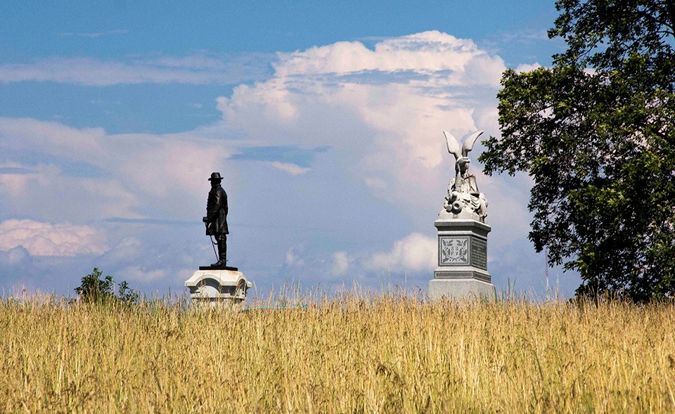 A blue sky with puffy white clouds can be seen behind two monuments at the horizon line with a wheat field in the foreground.