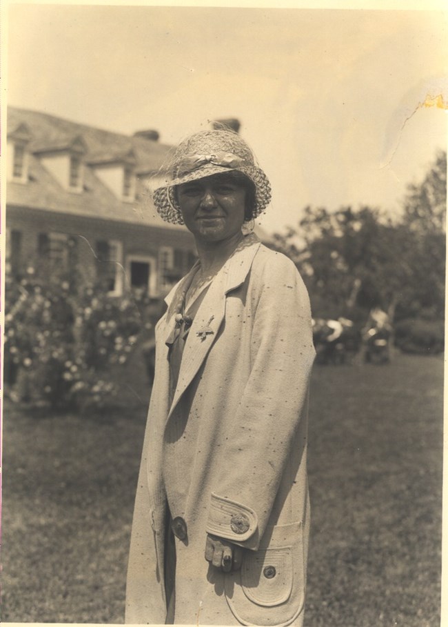 Old sepia photo of a woman in a hat and jacket standing in front of a brick building in the Memorial Area