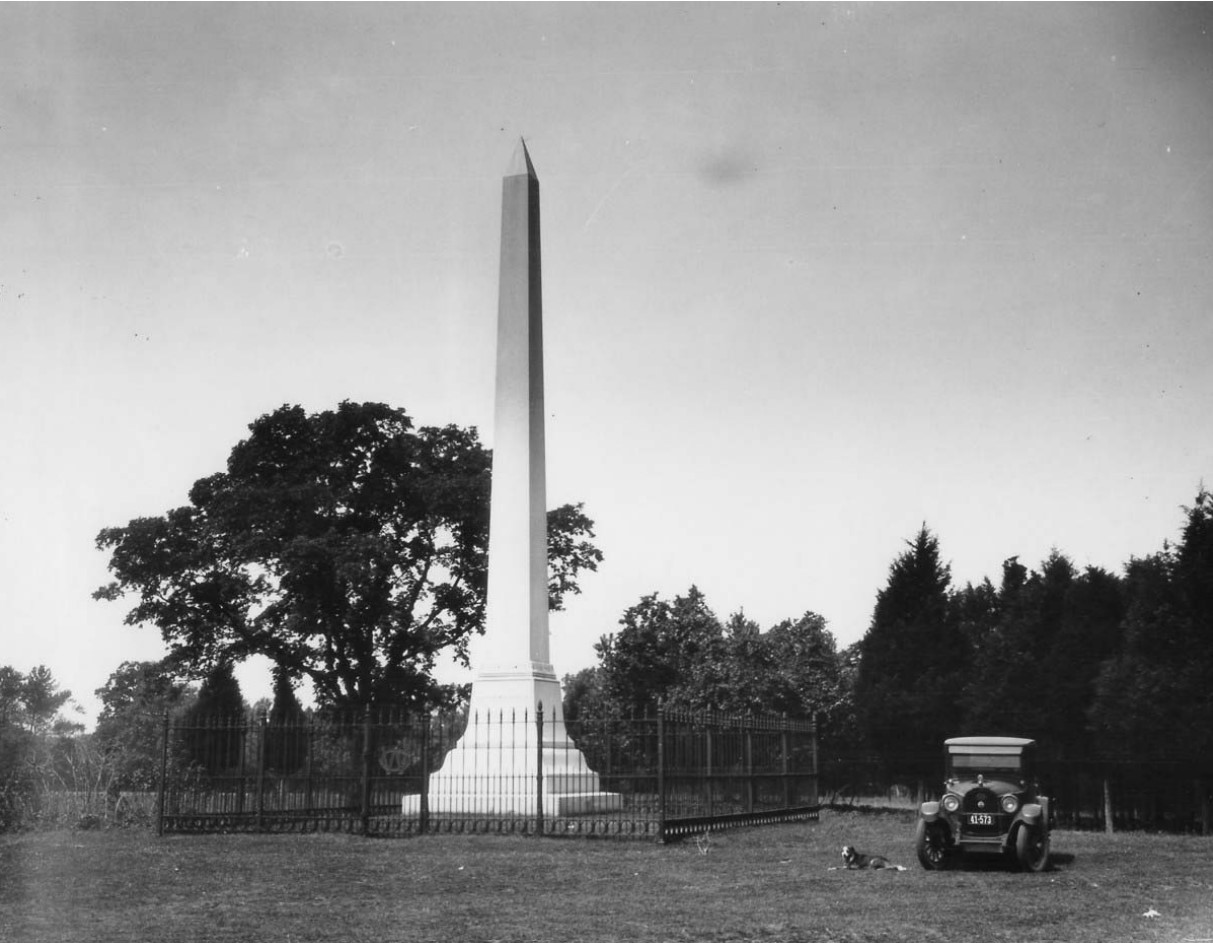black and white photo of the obelisk surrounded by a iron fence, a dog laying next to the fence and an old car parked to the side.
