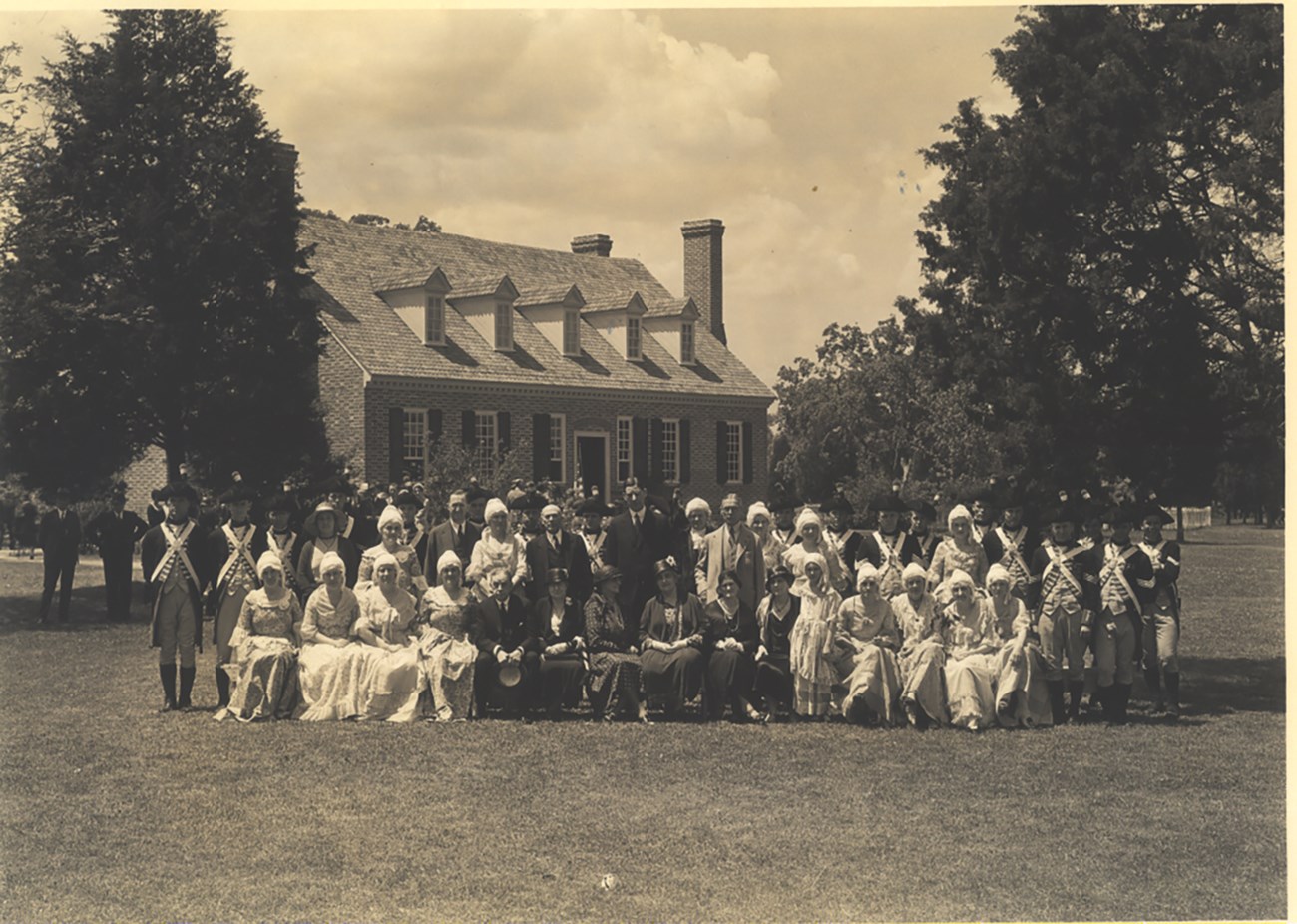 A sepia photo with a bunch of people in colonial garb sitting in front of the memorial house museum