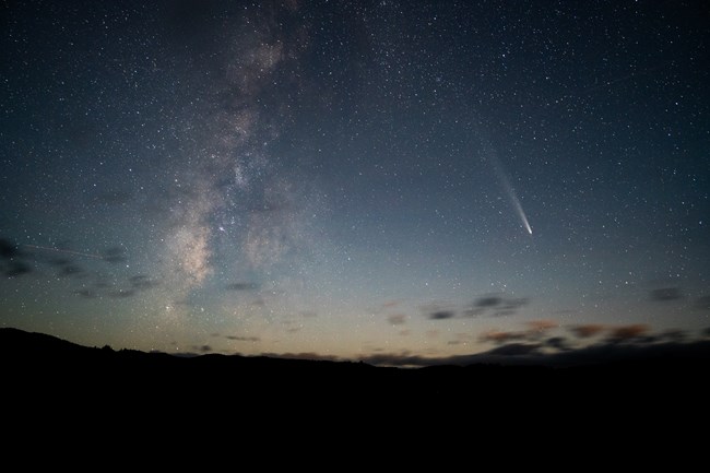 A bright comet with a glowing tail streaks across the night sky beside the hazy band of the Milky Way, both set against a field of countless stars.