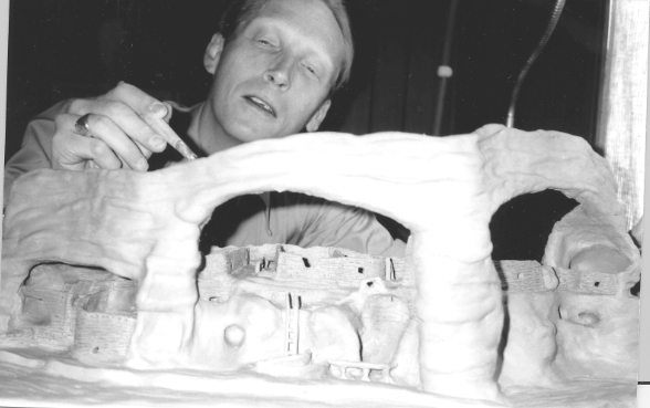 A model of 3 of the Cliff Dwellings' caves sits on a table as a man towers over it with paintbrush in hand, adding the final touches.