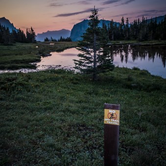 A small signs says, "No off trail travel," with a mountain sunrise in the background.