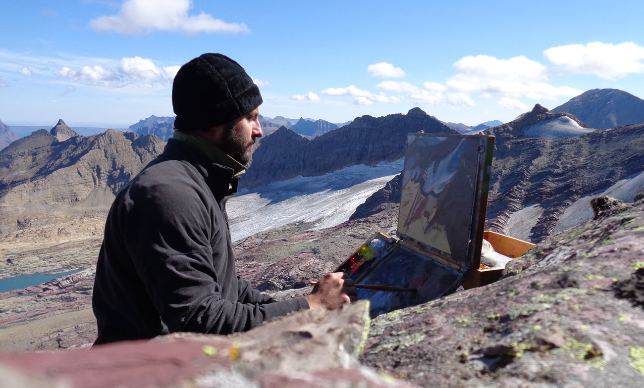 A man sits and paints a mountain landscape.
