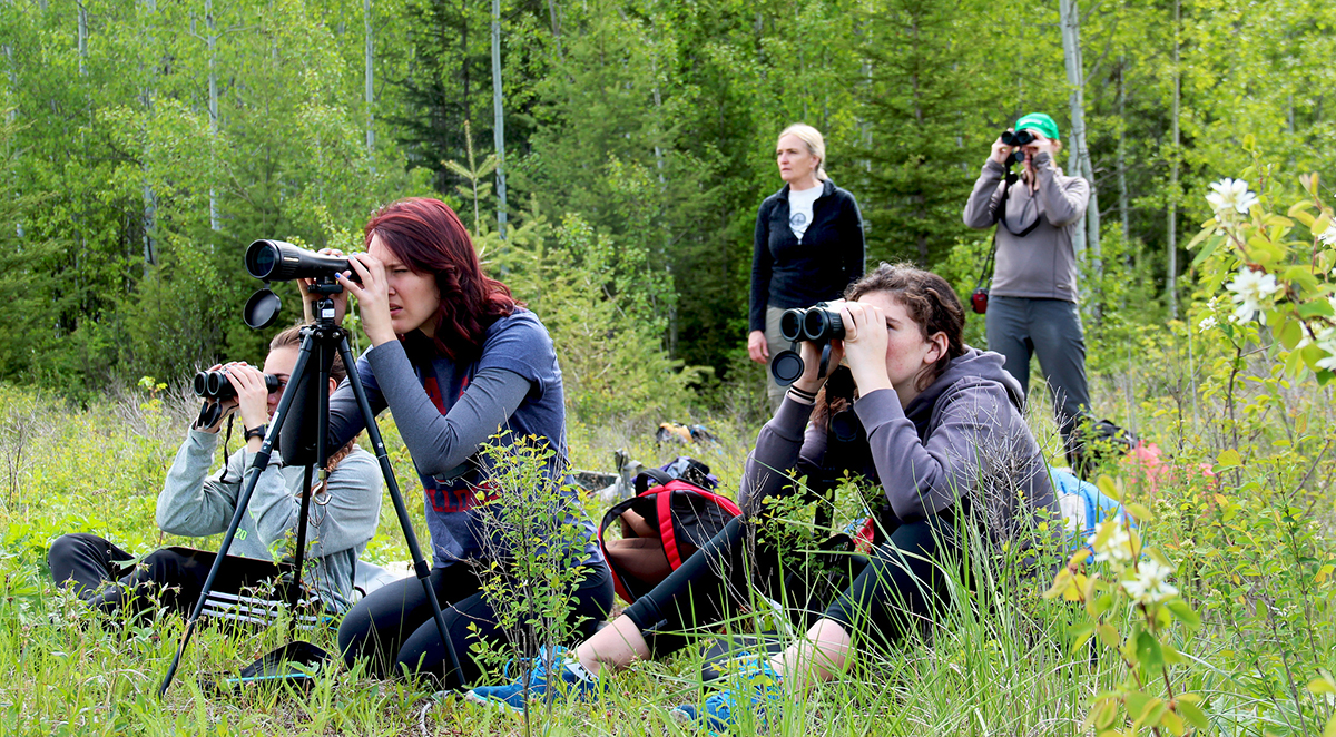 group of students look through binoculars while sitting on ground, with teachers standing behind