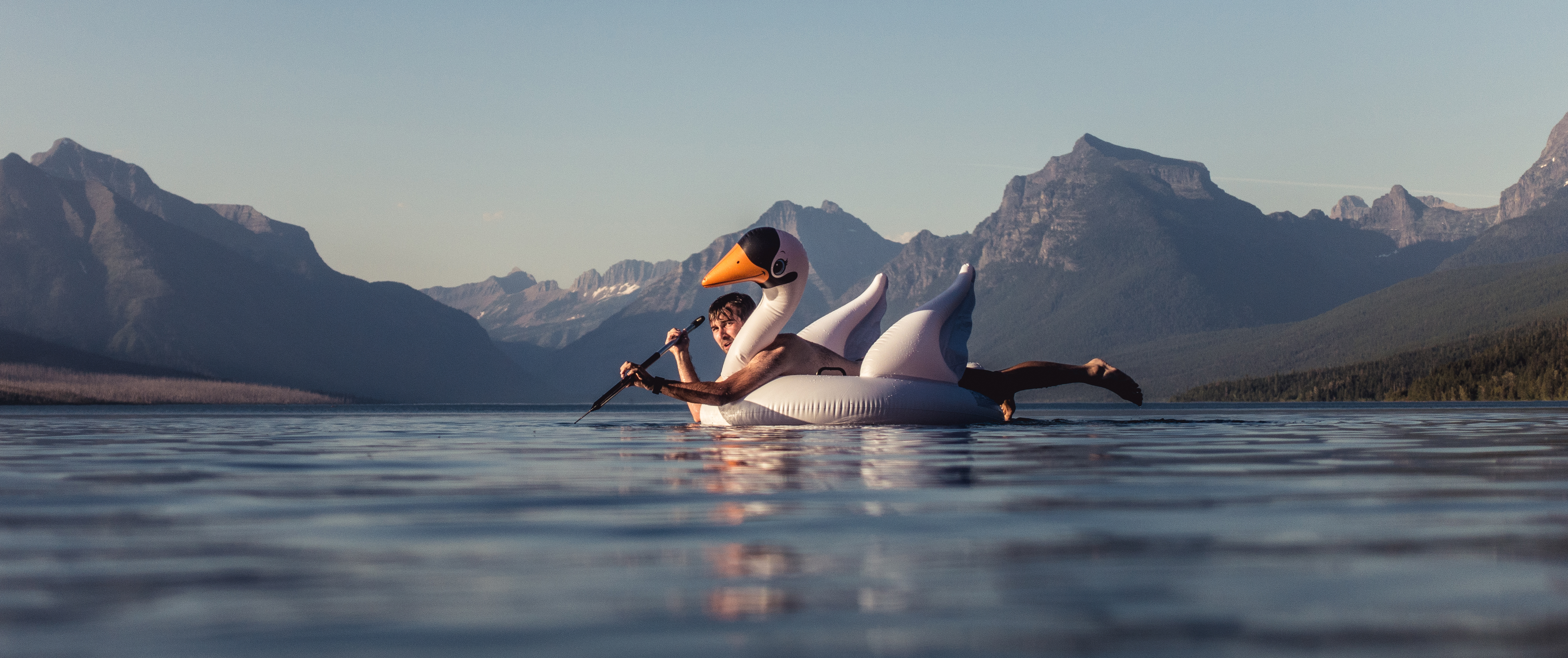 A man lays on a large inflatable swan while paddling across a mountain lake.
