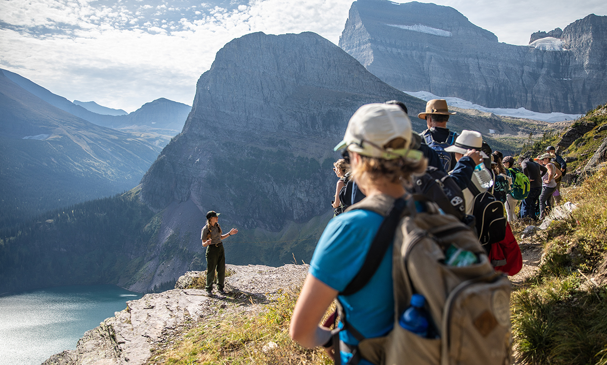 A ranger stands near the edge of a trail with mountains and a lake behind her while talking to a group of hikers.