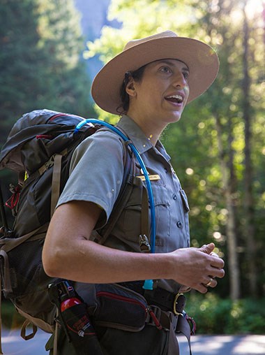 A woman wearing a National Park Service uniform and a backpack stands outside talking to visitors who are not in the image.
