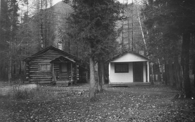 Black and white image of a log cabin on the left next to a white cabin on the right, both in a forest setting.