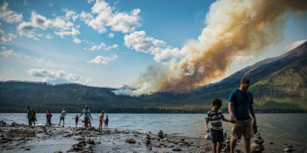 People along a lake shore with a wildland fire in the background.
