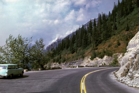 Photo taken from the middle of a road with a steep green hillside above it, with smoke billowing over a distant hillside. A 1960s car is parked in a pullout on the road.