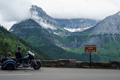 A motorcyclist parked at an overlook along a short stone wall. A waterfall from a deep "hanging" valley positioned above another trickles down in the distance.