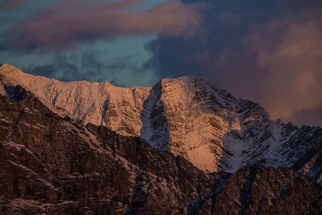 A mountain ridge covered in snow reveals layers of ridges that appear to have been folded and bent within the rock.