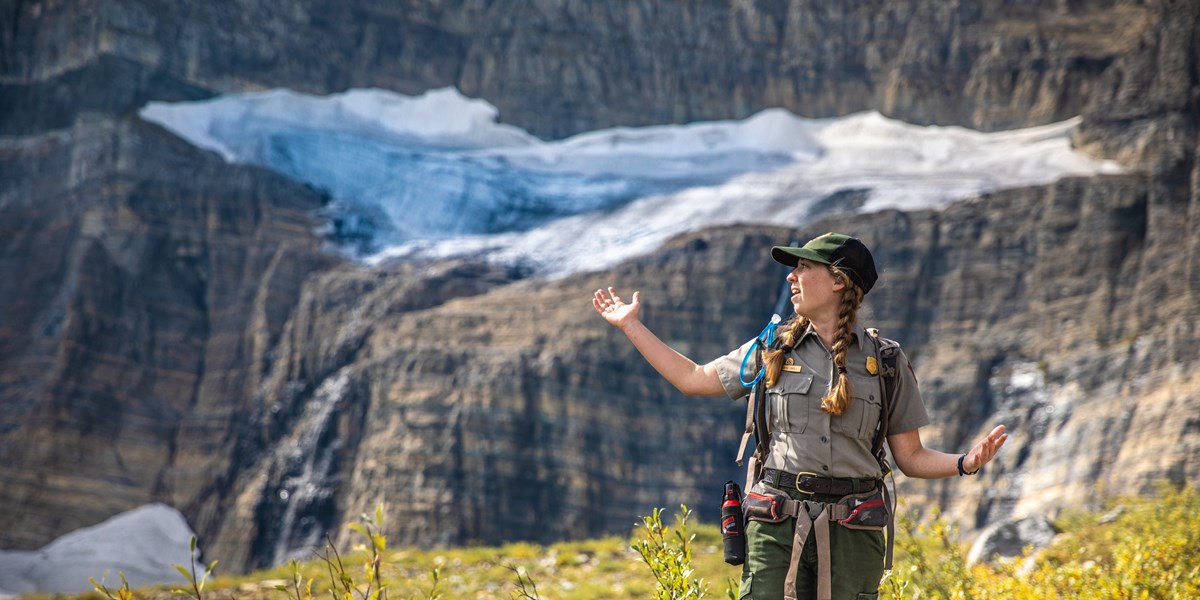A national park ranger stands with their arms out in front of a glacier.