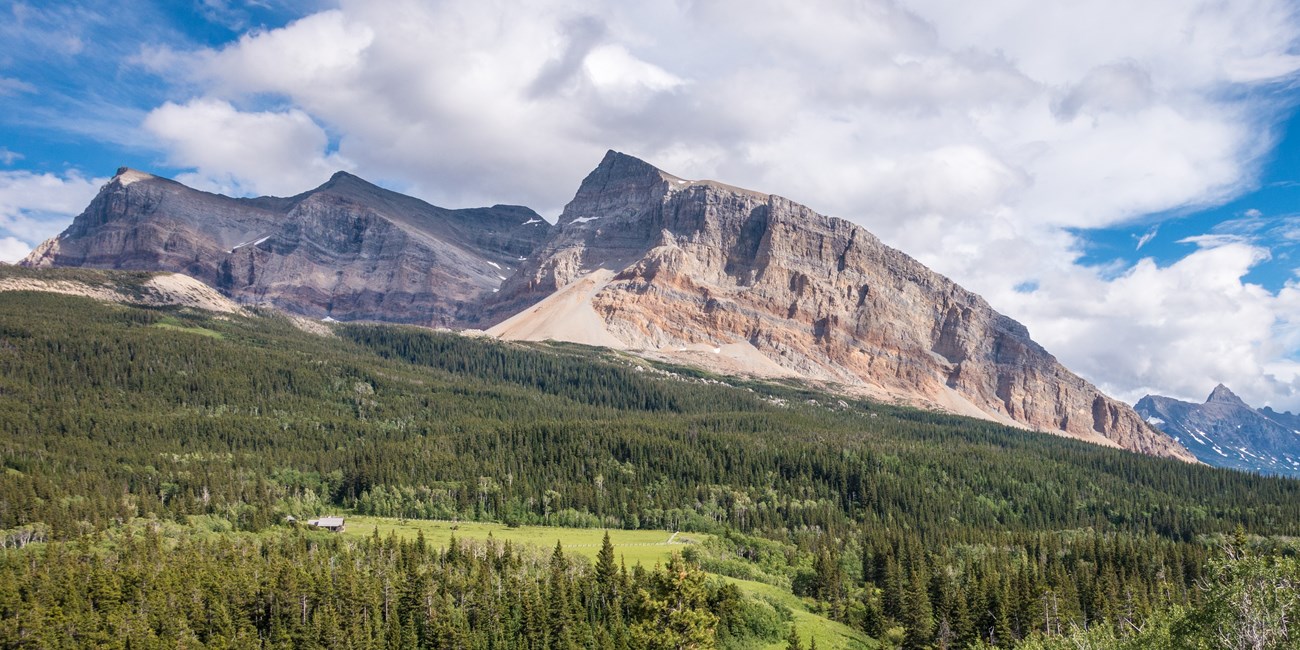 Mountains of pale brown rock ridge over a green hill of coniferous trees. A small cabin can be seen in the midst of a green clearing below.