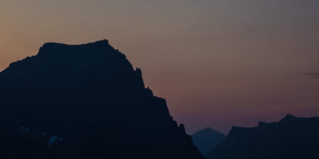 A silhouette of three mountains is seen with the backdrop of a darkening sky. It's clear of clouds and fades from orange to a dark purple.