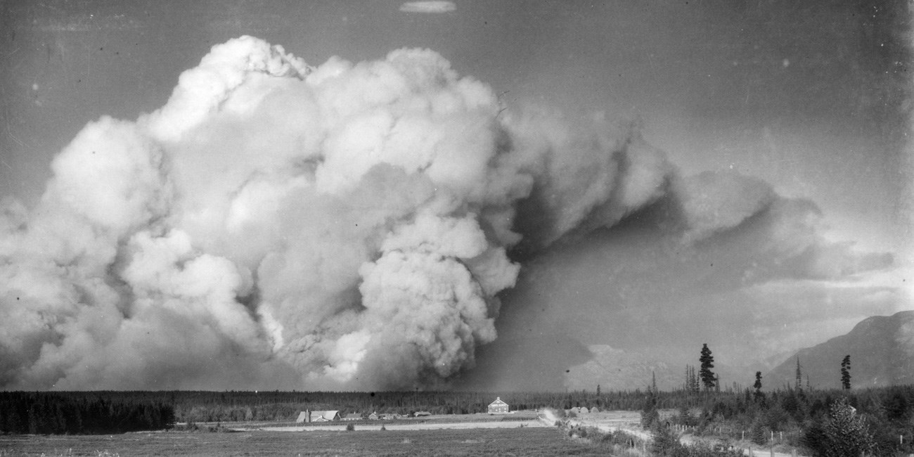 Black and white image of a billowing column of smoke dwarfing the view of the valley in the foreground and the mountains beyond.