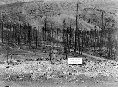 Black and white image of a pile of rubble with bits of masonry and metal, with a sign propped up that reads Many Glacier Ranger Station. It is surrounded by blackened trees and a steep hillside beyond.